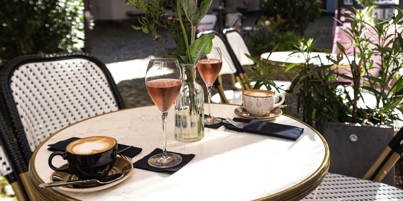 Gedeckter Cafétisch im Sonnenschein mit Cappuccinos, zwei Gläsern Roséwein und Blumen.