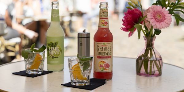 Zwei Flaschen Limonade (Limette-Minze und Pink Grapefruit) stehen neben passend garnierten Gläsern und einer Blumenvase auf einem weißen Tisch.