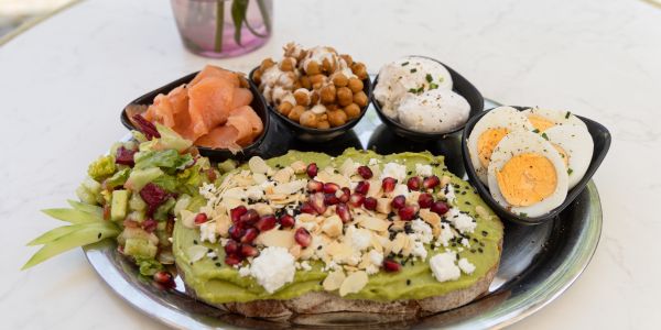 Eine Servierplatte mit Avocado-Schnittchen, Räucherlachs, Eiern, Kichererbsen und Salat steht angerichtet auf einem Tisch im Außenbereich.