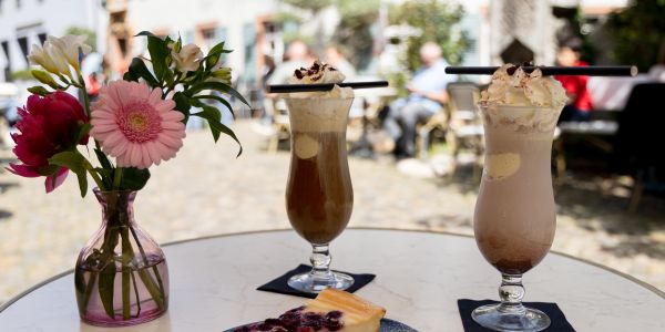 Ein Stück Beeren-Käsekuchen mit Puderzucker auf einem Teller vor zwei Gläsern Eiskaffee mit Schlagsahne stehen auf einem runden Tisch.
