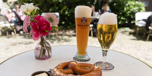 Eine Brezel mit Butter auf einem schwarzen Teller steht vor einem Glas Weißbier und einem Pils auf einem runden Cafétisch.