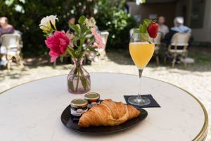 Ein Croissant auf einem Teller mit zwei kleinen Marmeladengläsern steht neben einem gelben Aperitif mit einer Erdbeere auf dem Tisch.