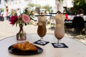 Ein Croissant auf einem schwarzen Teller neben zwei hohen Gläsern mit Eiskaffee und Schlagsahne sowie eine Blumenvase stehen auf einem runden Tisch.
