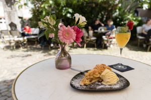 Ein Stück gedeckter Apfelkuchen mit Vanilleeis steht neben einem gelben Cocktail mit Erdbeere und einer bunten Blumenvase auf einem weißen Cafétisch.