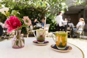 Zwei Gläser mit bunt geschichteten Matcha-Eistee-Kreationen (Erdbeere und Mango) stehen auf Untertellern neben einer Blumenvase im Außenbereich.