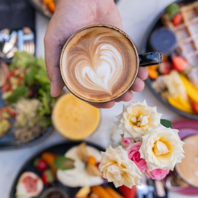 Eine Tasse Kaffe in der flachen Hand über einem reichhaltig gedeckten Tisch
