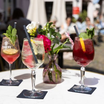 Drei sommerliche Spritz-Cocktails mit Strohhalmen und Minz-Garnitur auf Servietten stehen auf einem runden Tisch neben einer Blumenvase.