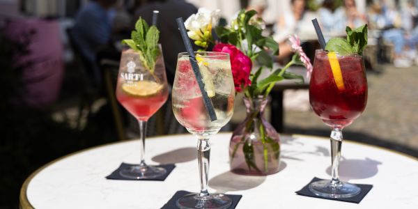 Drei sommerliche Spritz-Cocktails mit Strohhalmen und Minz-Garnitur auf Servietten stehen auf einem runden Tisch neben einer Blumenvase.