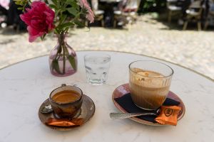 Eine Tasse Espresso und ein Glas Eiskaffee mit Keksen stehen serviert neben einem Glas Wasser und einer Blumenvase auf einem weißen Tisch.