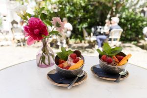 Zwei halbkugelförmige Metallschalen mit gemischtem Eis, frischen Beeren und Minze stehen auf Untertassen neben einer Blumenvase im Straßencafé.
